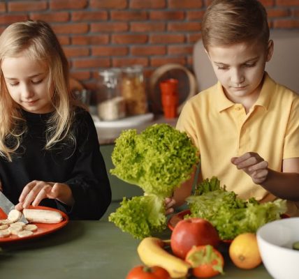 Focused little girl and boy sitting at desk with assorted vegetables and fruits while cooking in loft kitchen with brick wall